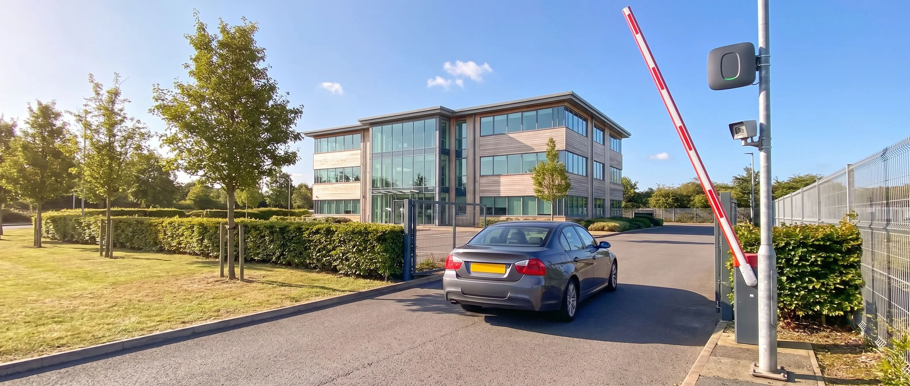 Dark grey saloon car driving through the raised barrier at a business park entrance, with a uPASS Go reader and a camera on the post and a modern office building beyond the landscaped verge