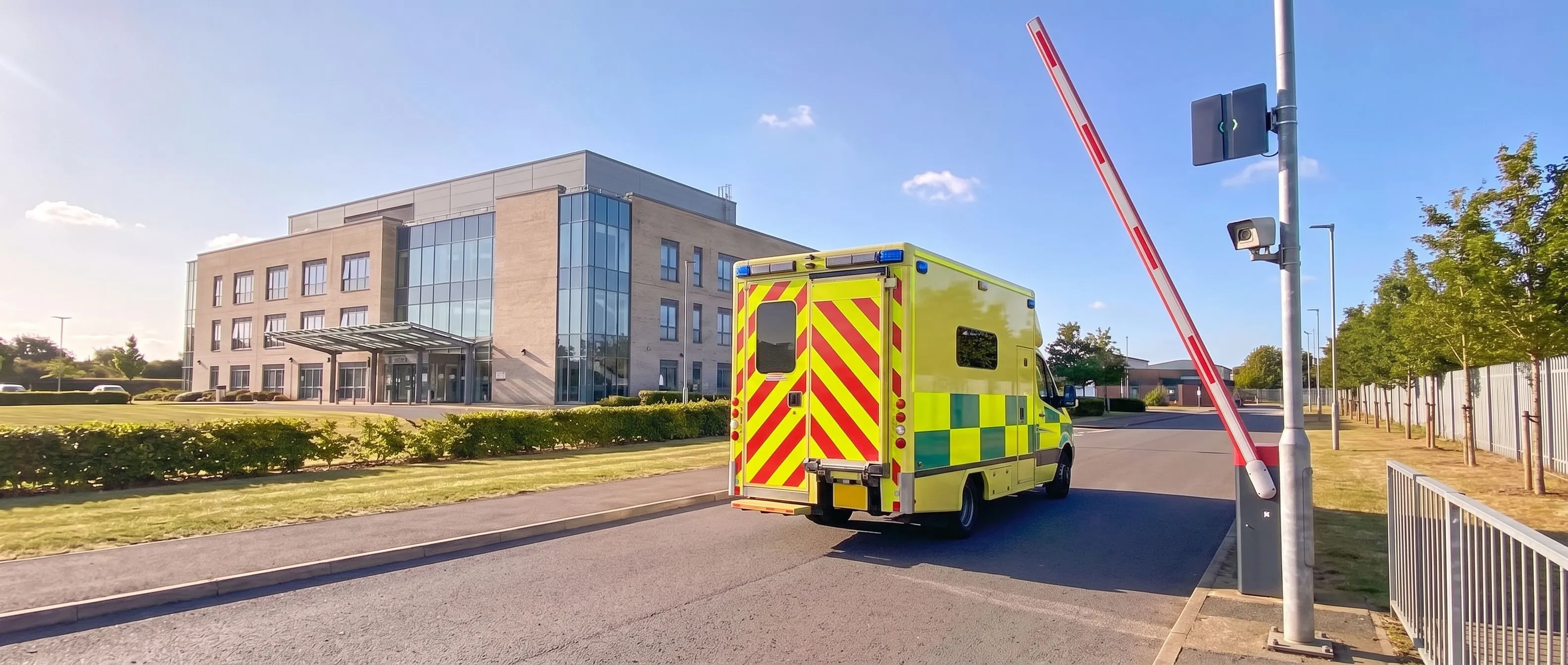 Yellow and green ambulance driving through the raised barrier at a hospital's perimeter gate towards the main building, with a Transit Ultimate long-range reader and a camera on the post beside the lane