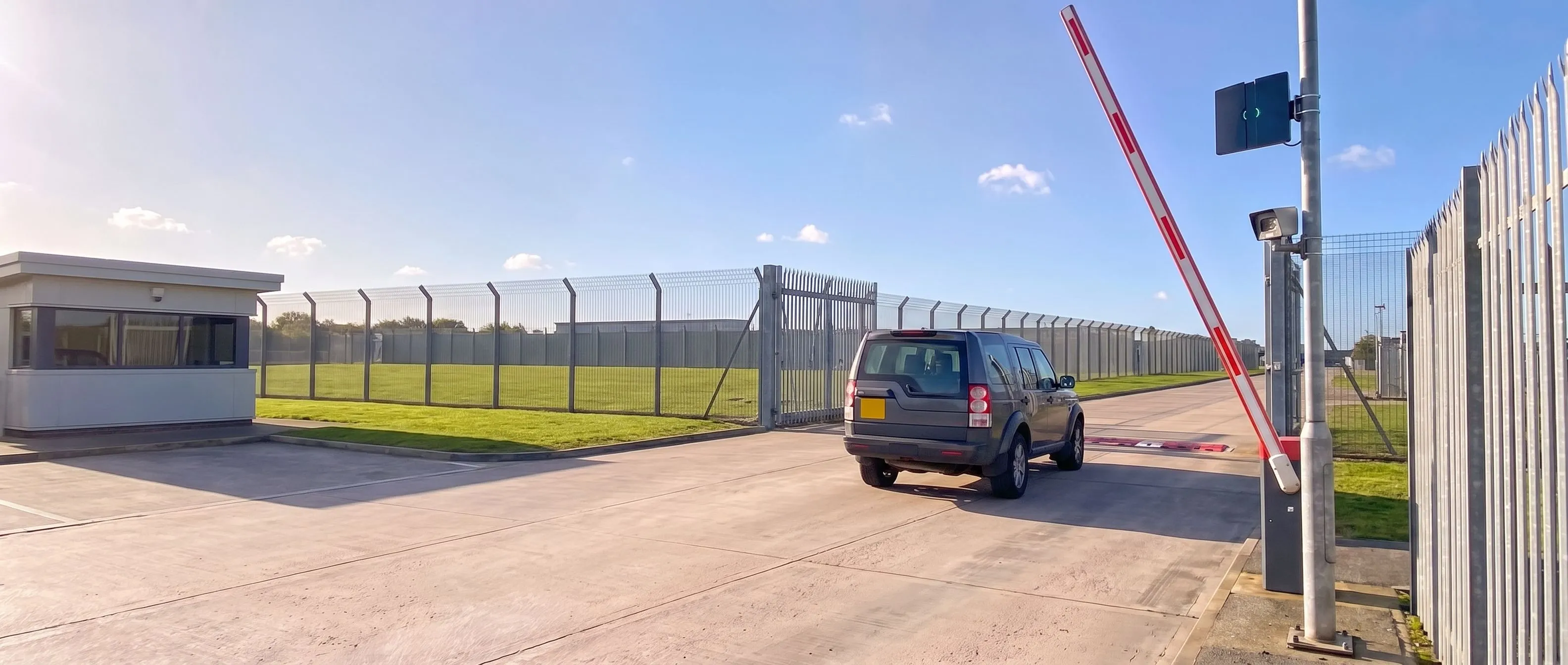 Grey 4x4 driving through the raised barrier at a secure estate gate, with a lowered road blocker in the lane, a Transit Ultimate long-range reader and a camera on the post, a guardroom and weldmesh perimeter fencing